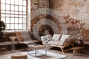 Armchair and beige sofa in industrial living room interior with wooden table, pouf and window