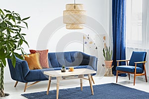 Armchair and sofa in blue and orange living room interior with lamp above wooden table. Real photo