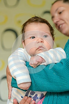 In the arms of a cheerful mom. Portrait of charming toddler with blue eyes and chubby cheeks resting vertically in his mother`s