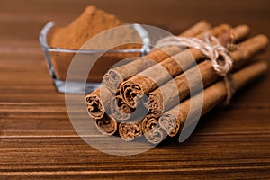 Aromatic cinnamon sticks and glass bowl with powder on wooden table, closeup