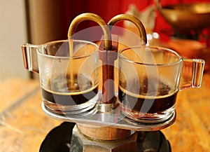 Aromatic coffee trickling out from mini retro brewing pot into a pair of glass demitasse cups