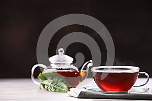 Aromatic hot tea in glass cup, teapot and leaves on white wooden table against black background. Space for text