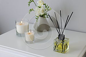 Aromatic reed air freshener, freesia flowers and candles on white bedside table indoors