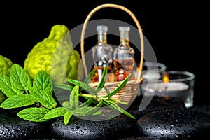 Aromatic spa of bottles essential oil in basket, fresh mint, rosemary, bergamot fruits and candles on black zen basalt stones