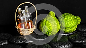 Aromatic spa concept of bottles essential oil in basket, bergamot and zen basalt stones with dew on black background, closeup