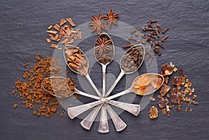 Aromatic spices and brown sugar in silver spoons on black stone background