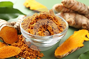 Aromatic turmeric powder and raw roots on green background, closeup