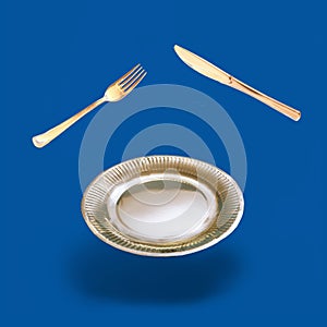 Arrangement made of levitating golden color fork, knife and plate. Dark blue background