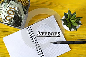 ARREARS - written in a notebook on a yellow wooden background with a cactus and a stationery basket with crumpled bills