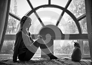 Art photo blond girl and white cat sitting at big old window during the rain. Romantic Black and white photo, loneliness