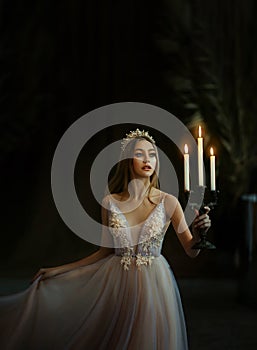 Art photo of medieval girl princess walks in dark gothic room. Woman queen is holding candlestick with burning candles