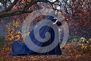 Art photography. Woman witch holds book in hands reads spell. Black clothes, cloak dress flies in wind. Fantasy girl elf