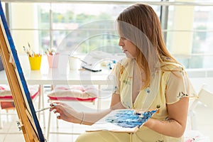 Art school, creativity and leisure concept - student girl or young woman artist with easel, palette and paint brush