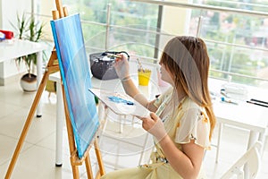 Art school, creativity and leisure concept - student girl or young woman artist with easel, palette and paint brush