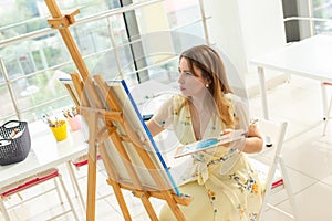 Art school, creativity and leisure concept - student girl or young woman artist with easel, palette and paint brush