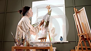 Art studio - young pretty woman in stained white shirt changing brushes and drawing a painting