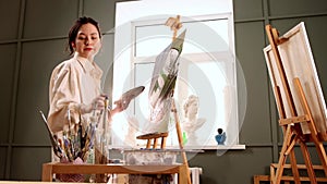 Art studio - young woman in stained white shirt changing brushes and drawing a painting