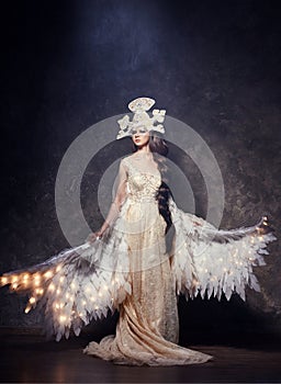 Art Woman angel with wings in luxurious long dress and fabulous headpiece. Girl bird with luminous wings posing on dark background