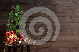 artificial christmas tree with red baubles, gift box and decoration on an small table in front of a rustic wooden wall, generous