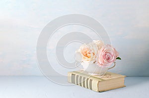 Artificial pink rose flowers in white cup with old book on blue background