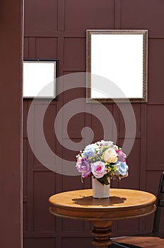Artificial rose flowers in ceramic vase on wooden round table with teak wood chair and the old blank picture frame on wooden wall
