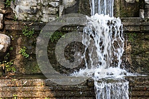 An artificial three-level waterfall flowing against the background of a stone`s overgrown moss wall.