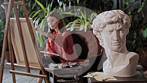 Artist in an art studio with plants - young pretty woman drawing a painting - greek head sculpture on a foreground