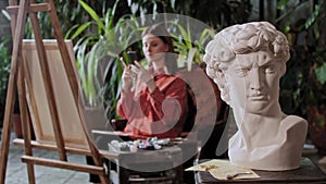 Artist in an art studio with plants - young pretty woman takes a photo of her painting - greek head sculpture on a