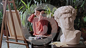 Artist in an art studio with plants - young woman artist put the canvas away from an easel - greek head sculpture on a