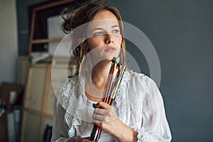 An artist female holding a pinch of brushes in her art studio. A young woman student painter searching for imagination at home