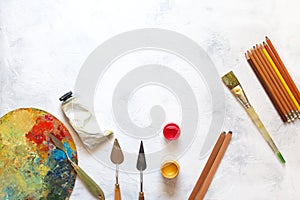 An artist`s palette covered with paint, painting knives, white paint tube, painting brush and pencils on a white background