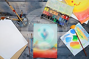 The artistÃ¢â¬â¢s supplies and his hand-made watercolor are laid out on a gray wooden table. On the table are open watercolor paints photo