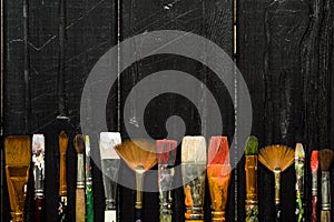 Artist`s tools, brushes in paint are in a row on a black wooden background.