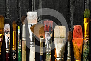 Artist`s tools, brushes in paint are in a row on a black wooden background.