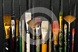 Artist`s tools, brushes in paint are in a row on a black wooden background.