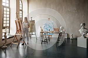 Artist at the working process. Young woman creating the painting. Workshop room with light and classical sculpture busts