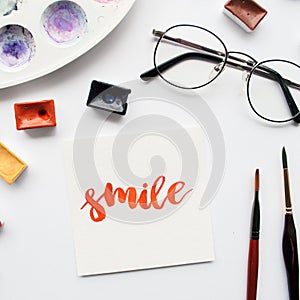 Artist workspace. Word Smile written in calligraphy style, watercolor cuvettes and palette on a white background.