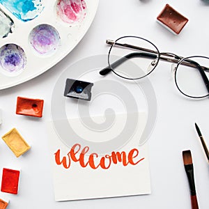 Artist workspace. Word Welcome written in calligraphy style, watercolor cuvettes and palette on a white background.