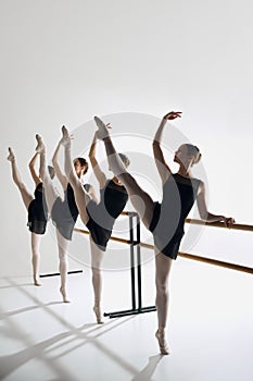 Artistic ballet pose. Four girls, ballet dancers in studio standing at barre, reaching out gracefully with one arm