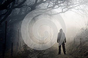 An artistic double exposure of a lone hooded figure on a path in the countryside on a spooky, foggy winters day.