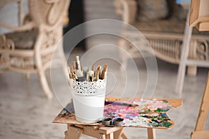 Artistic equipment: easel, paint brushes, tubes of paint, palette and paintings on work table in a artist studio.