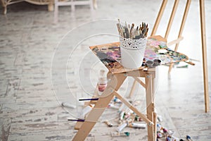 Artistic equipment: easel, paint brushes, tubes of paint, palette and paintings on work table in a artist studio.