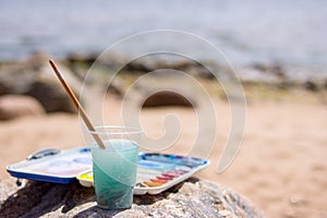 Artistic equipment: paint brushes, tubes of paint, palette and paintings on rock in nature sea shore at sunny summer day - creatio