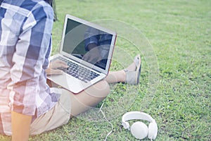 Asain woman wearing a shirt sitting on the green grass while typing Thai keyboard of laptop put on the leg with the ear plug for r