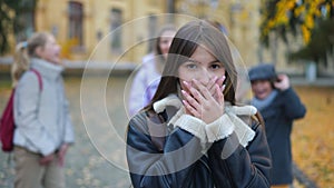 Ashamed bullied Caucasian teen girl covering mouth with hands looking at camera as blurred classmates laughing out loud