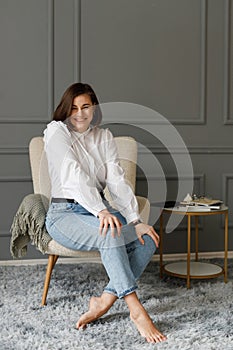 Ashamed smiling business girl wearing a white shirt and jeans with a wristwatch on her hand sitting in a beige chair with a gray