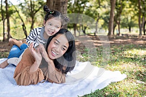 Asia daughter riding mother back and lying on the ground in the public garden under sunlight with copy space. The cute girl is