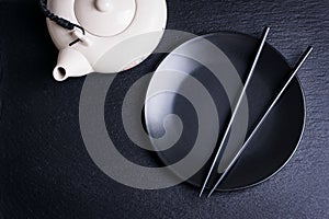 Asia food composition with chinese chopsticks, teapot and empty plate on a dark stone background