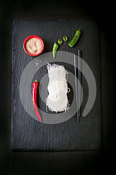 Asia food - rice noodles, spicy pepper, sticks and sesam, ready to cook on a dark stone background