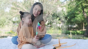 Asia mother and daughter sitting in the garden. Kid blowing colorful of plastic pinwheel and having fun in summer season. Family a